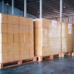 Stacked corrugated shipping boxes on pallets inside a modern warehouse packaging supplier facility.